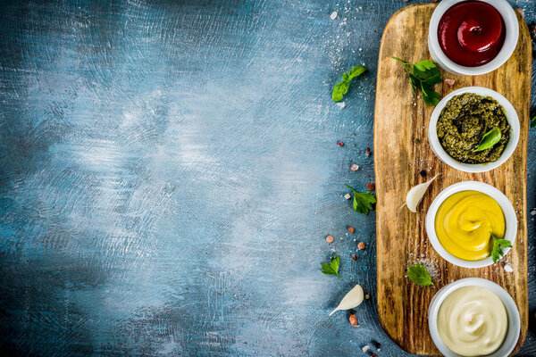 Set of four classic sauces - ketchup, mayonnaise, mustard, pesto - with herbs and spices. Blue concrete background top view copy space