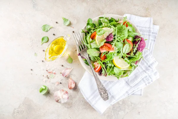 Fresh spring detox mix salad with vegetables (cucumber, lemon, tomato, arugula, and baby spinach), on light slate, stone or concrete background. Top view copy space