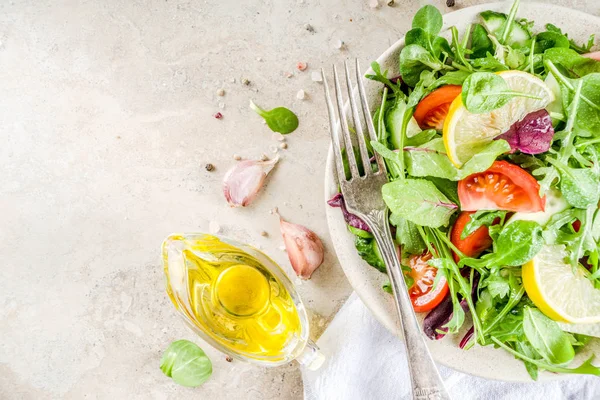 Fresh spring detox mix salad with vegetables (cucumber, lemon, tomato, arugula, and baby spinach), on light slate, stone or concrete background. Top view copy space