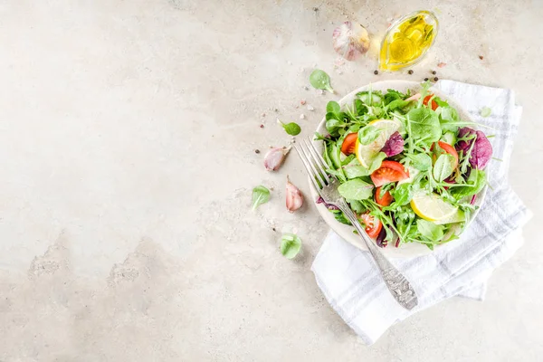 Fresh spring detox mix salad with vegetables (cucumber, lemon, tomato, arugula, and baby spinach), on light slate, stone or concrete background. Top view copy space