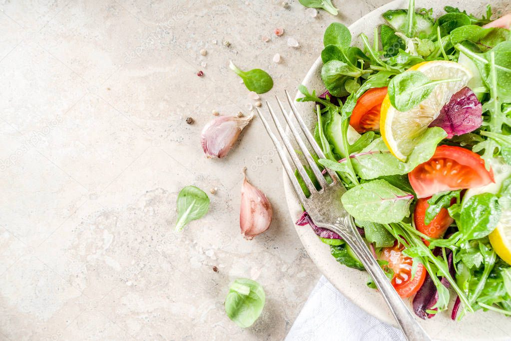 Fresh spring detox mix salad with vegetables (cucumber, lemon, tomato, arugula, and baby spinach), on light slate, stone or concrete background. Top view copy space