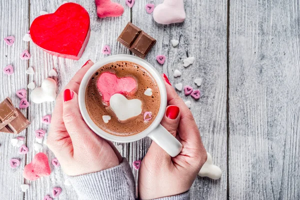 Girl hands hold hot chocolate with marshmallow hearts, red pink white color with chocolate pieces, sugar sprinkles, old wooden background copy space top view, hands in pictute flatlay