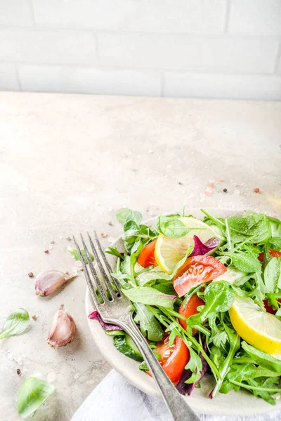 Fresh spring detox mix salad with vegetables (cucumber, lemon, tomato, arugula, and baby spinach), on light slate, stone or concrete background. Top view copy space