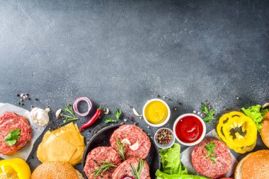 Hamburger arka planı. Çeşitli cheeseburger ve sığır burger malzemelerini hazırla - çörek, domates, soğan, marul, sos, peynir ve çiğ burger pirzolası, ızgara için hazır. Burger bbq parti festivali konsepti, üst görünüm kopyalama alanı