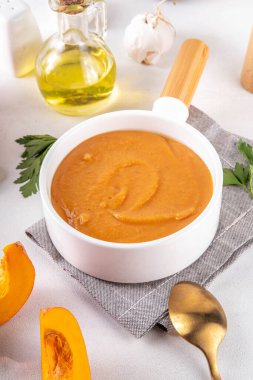 Beyaz kasede servis edilen balkabağı kremalı çorba. Geleneksel sonbahar ve kış yemekleri, sıcak ve baharatlı balkabağı çorbası, kremalı ve lezzetli, balkabağı dilimleri ve beyaz masa üzerinde bitkiler