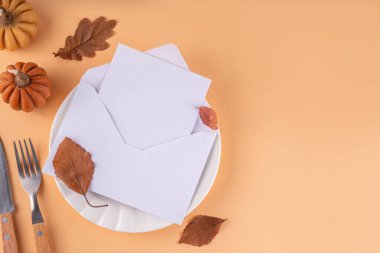 Sonbahar sofrası kurma, küçük balkabaklarıyla parti davetiyesi, güz yaprakları, tabak, çatal bıçak takımı ve bir davetiye veya selamlama ile birlikte zarf. Şükran Günü partisinin rahat arka plan fotokopi alanı