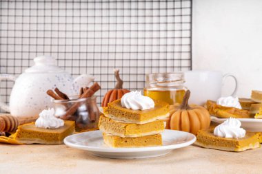 Homemade autumn taste pumpkin pie bars. Freshly baked pumpkin bars with whipped cream on it, with autumn decor and spices on beige table background, copy space 