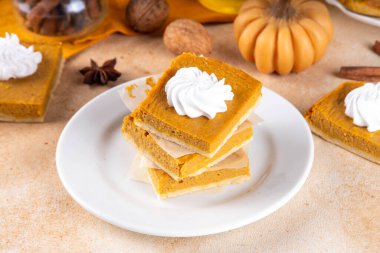 Homemade autumn taste pumpkin pie bars. Freshly baked pumpkin bars with whipped cream on it, with autumn decor and spices on beige table background, copy space 