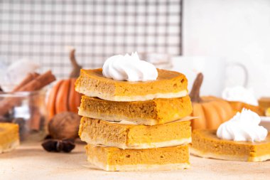 Homemade autumn taste pumpkin pie bars. Freshly baked pumpkin bars with whipped cream on it, with autumn decor and spices on beige table background, copy space 