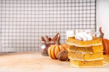 Homemade autumn taste pumpkin pie bars. Freshly baked pumpkin bars with whipped cream on it, with autumn decor and spices on beige table background, copy space 