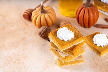 Homemade autumn taste pumpkin pie bars. Freshly baked pumpkin bars with whipped cream on it, with autumn decor and spices on beige table background, copy space 