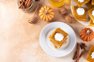 Homemade autumn taste pumpkin pie bars. Freshly baked pumpkin bars with whipped cream on it, with autumn decor and spices on beige table background, copy space 