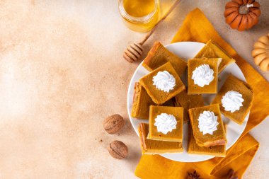 Homemade autumn taste pumpkin pie bars. Freshly baked pumpkin bars with whipped cream on it, with autumn decor and spices on beige table background, copy space 