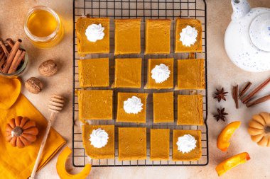 Homemade autumn taste pumpkin pie bars. Freshly baked pumpkin bars with whipped cream on it, with autumn decor and spices on beige table background, copy space 