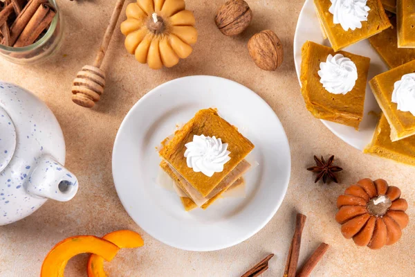 Homemade autumn taste pumpkin pie bars. Freshly baked pumpkin bars with whipped cream on it, with autumn decor and spices on beige table background, copy space 