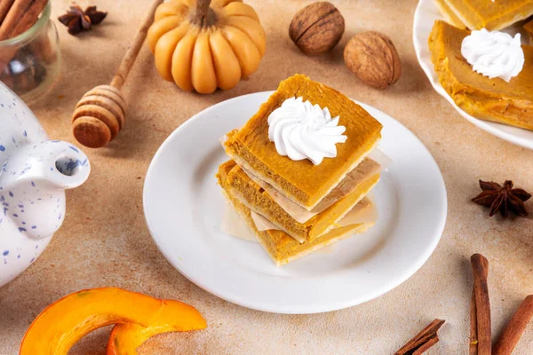 Homemade autumn taste pumpkin pie bars. Freshly baked pumpkin bars with whipped cream on it, with autumn decor and spices on beige table background, copy space 