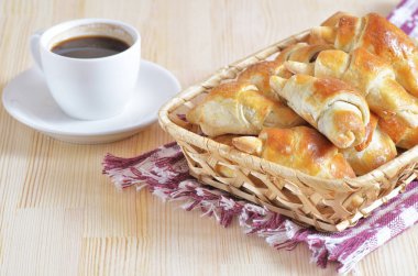 Yanındaki beyaz fincan kahve masada bir masa örtüsü üzerinde bir hasır sepet içinde ev yapımı ay çöreği.