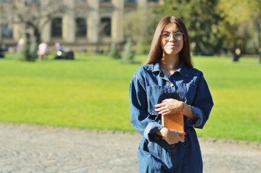 Üniversite parkının arka planında gözlüklü bir kitapla duran güzel, mutlu genç bir öğrenci.