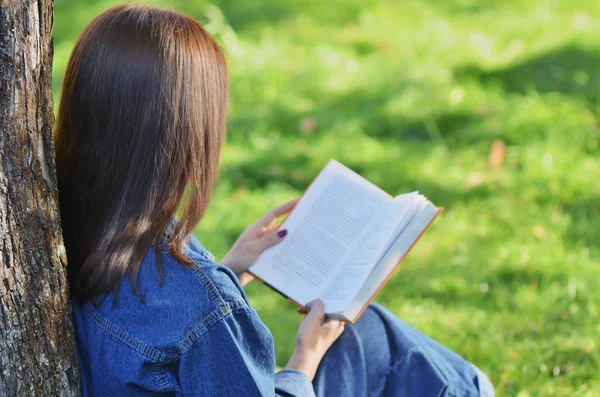 Güzel, mutlu, kitap okuyan genç bir kız kampüs, üniversite, okul, eğitim yakınlarındaki bir ağacın altında yeşil çimlerde oturuyor. Yaz, bahar, yeşil Park, parlak güneş