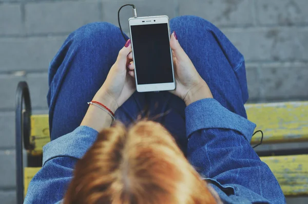 Genç ve güzel bir öğrenci, sokakta sarı bir bankta oturan bir akıllı telefon kullanıyor.