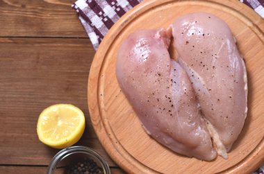 Çiğ tavuk göğsüne karabiber ve tuz serpiştirilmiş. Yuvarlak ahşap bir tahtanın üzerinde, bir limonun yanında. Yatay yakın plan fotoğraf