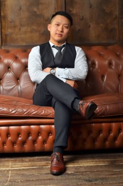 Başarılı bir Asyalı adam deri koltukta oturur, ayağını bacağına koyar ve ellerini çaprazlar.