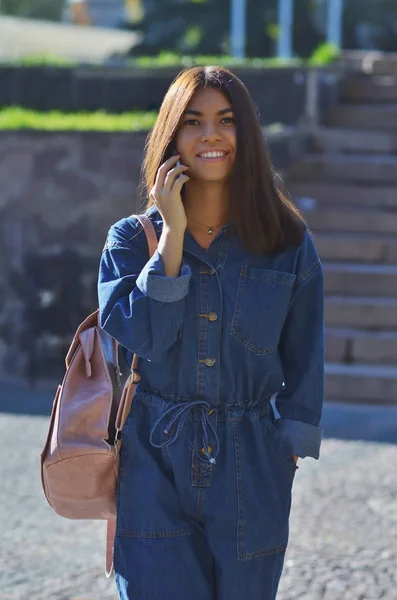 Una joven sonríe, camina por la ciudad con una mochila rosa hablando por teléfono —  Fotos de Stock