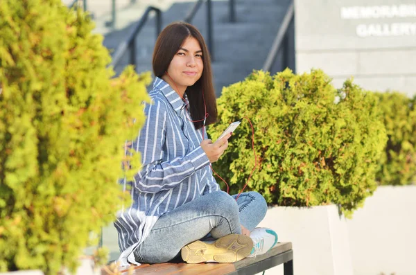 Joven hermosa es asiática chica se sienta en el banco cerca de verde tress y escuchar música —  Fotos de Stock