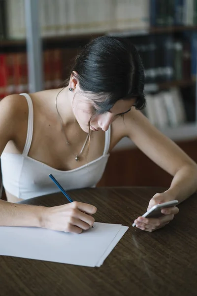 一个年轻女子的背部视图在一个图书馆与智能手机的桌子上学习 — 图库照片