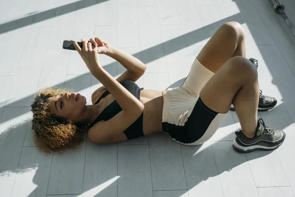 Γυναίκα φορώντας ένα αθλητικό σουτιέν και ένα κοντό τζιν ξαπλωτικό στο πάτωμα. Κρατώντας το τηλέφωνο — Φωτογραφία Αρχείου