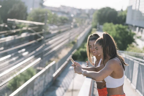 Δύο αρκετά αθλητικές φίλες κοιτάζοντας το smartphone και αγκαλιάζοντας στο δρόμο. — Φωτογραφία Αρχείου