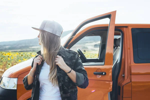 Çekici sarışın kız turuncu minibüs kapısında ağaçlar ve mavi gökyüzüyle poz veriyor.