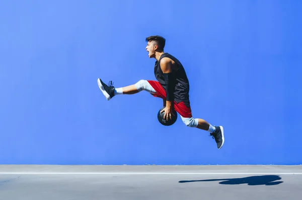 Zijaanzicht van een jongeman met een basketbal die voor een blauwe muur springt. — Stockfoto