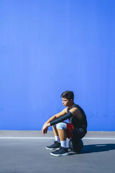 Side view of a man sitting on basket ball in front of a blue wall. — Stock Photo, Image