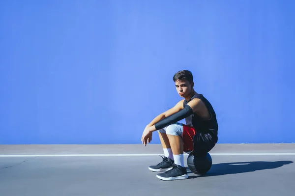 Vista lateral de um homem sentado na bola da cesta na frente de uma parede azul . — Fotografia de Stock