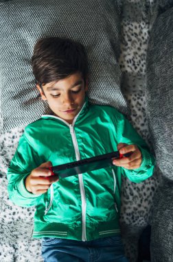 Çocuk kanepede uzanıp evdeki bilgisayar oyunu konsoluyla oynuyor.