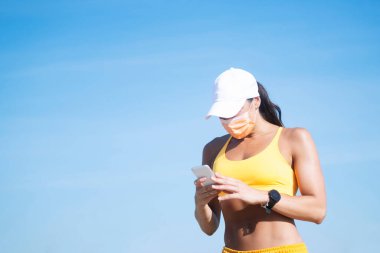 Atletik kadın dışarıda yüz maskesi takan akıllı telefonunu kullanıyor.