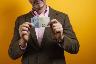 Elinde 20 Euro 'luk banknot tutan kahverengi ceketli ve pembe gömlekli bir adamın yakın çekimi.