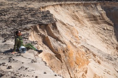 Yeşil bir uzay elbisesi içinde bir astronot kumlu bir uçurumda oturuyor ve çorak bir çöl manzarasına bakıyor..