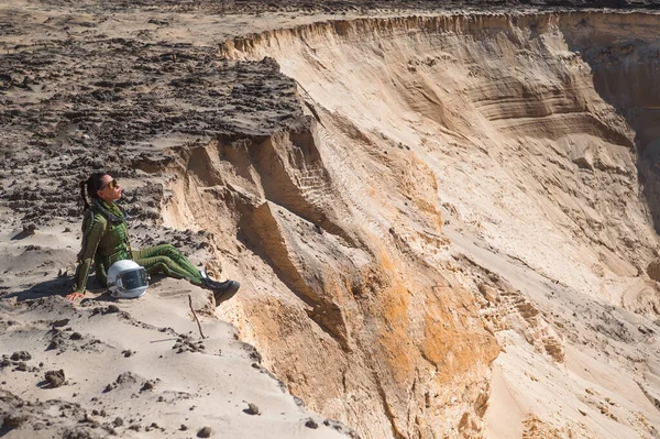 Yeşil bir uzay elbisesi içinde bir astronot kumlu bir uçurumda oturuyor ve çorak bir çöl manzarasına bakıyor..
