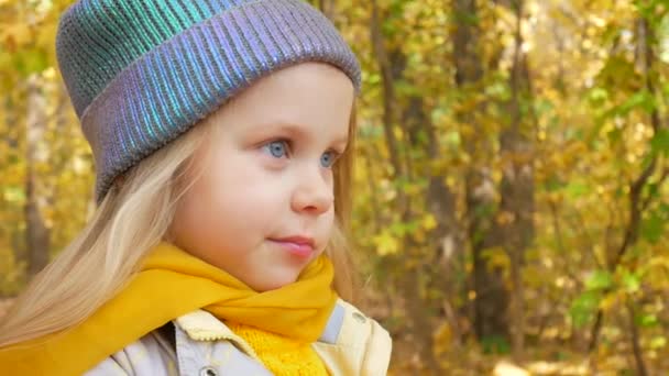 Niña linda en azul sombrero de color neón y bufanda amarilla está sonriendo en el parque en otoño — Vídeo de stock