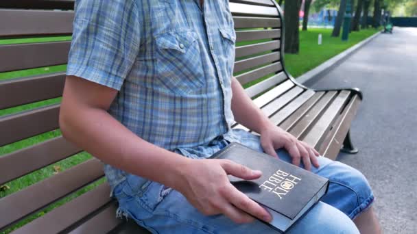 El hombre sostiene la Santa Biblia en su mano sentado en un banco en un parque en el verano en un día soleado — Vídeos de Stock