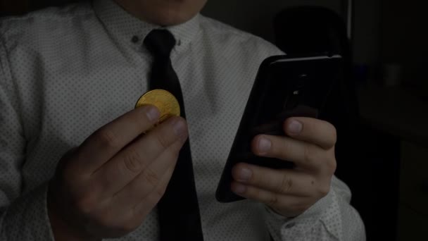 Trader in white shirt with tie using black smartphone and holds in hand golden bitcoin BTC coin — 비디오