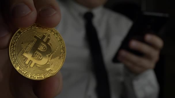 Trader en chemise blanche avec cravate à l'aide d'un smartphone noir et tient dans la main Bitcoin d'or pièce BTC — Video