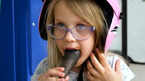 Kleines Mädchen mit blonden Haaren und schmutzigem Mund in rosa Motorradhelm und Brille isst Schokoladeneis Waffelkegel in der Straße