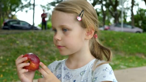 Klein meisje eet rode appel op groene park achtergrond — Stockvideo
