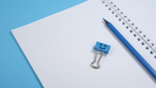 Bloc de notas de papel en blanco con lápices azules y clip de carpeta de sonrisa sobre fondo azul — Vídeos de Stock