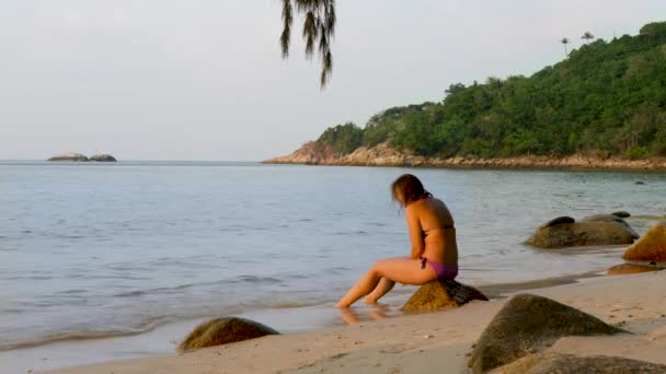 身穿紫色泳衣的漂亮女子坐在海滩上的石头上 — 图库视频影像