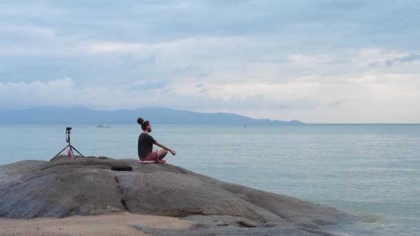 Ung man med dreadlocks mediterar i Lotus Pose On Rock By Sea och registrerar sig med kameran — Stockvideo