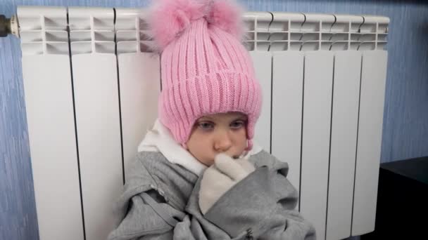 Menina pequena desgaste em roupas quentes e chapéu de malha congelar em casa senta-se perto do sistema de aquecimento do tubo central — Vídeo de Stock